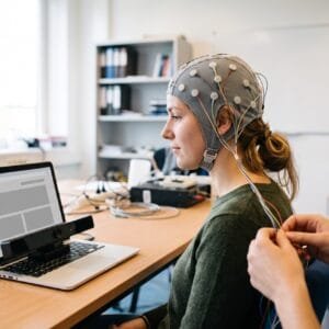 Jonge vrouw met een EEG-kap kijkt naar een laptop met een eye tracker eronder in een onderzoeksruimte