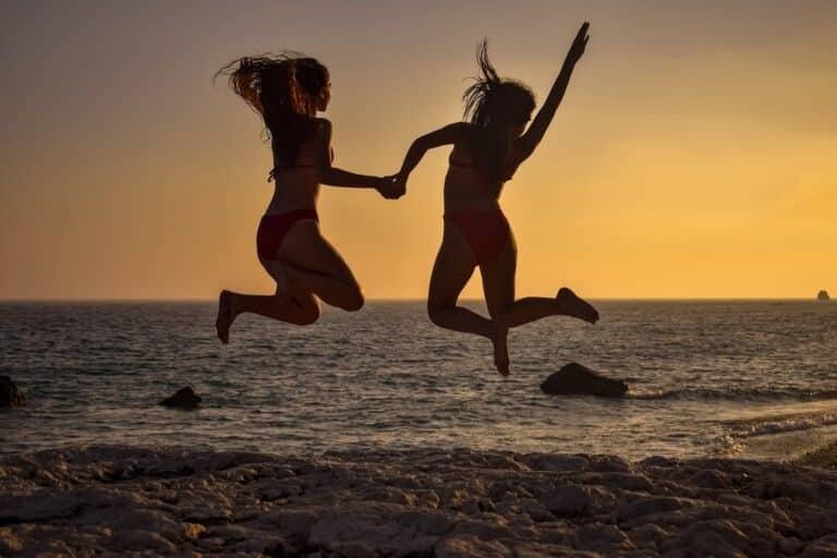 Twee vrouwen springen hand in hand op het strand bij zonsondergang