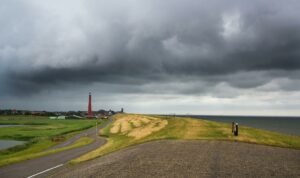 Dijk langs het water met een vuurtoren in de verte onder donkere wolken