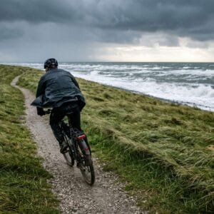 Fietser op een zeedijk bij harde wind, met schuimende golven en donkere wolken boven de Noordzee