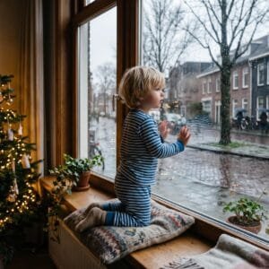 Kind in pyjama kijkt op een grijze decemberochtend door het raam naar een natte groene straat zonder sneeuw