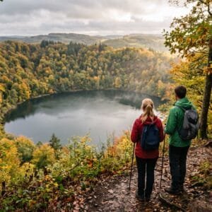 Wandelaars kijken vanaf de beboste kraterrand neer op een rond kratermeer in de Eifel in de herfst