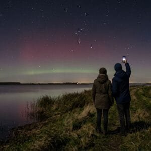 Twee mensen op een dijk bij het Lauwersmeer kijken in de nacht naar een rode gloed van noorderlicht in het noorden