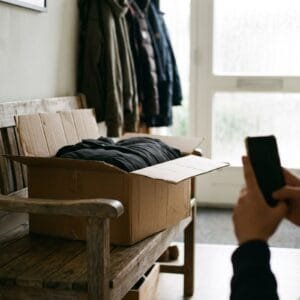 Handen houden een telefoon vast bij een geopend pakket met een opgevouwen jas op een bankje in de hal
