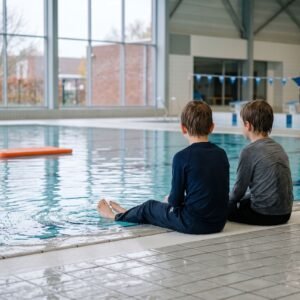 Twee kinderen zitten met kleren aan op de rand van een binnenzwembad, van achteren gezien