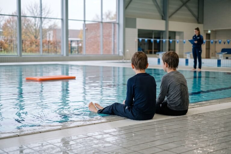 Twee kinderen zitten met kleren aan op de rand van een binnenzwembad, van achteren gezien