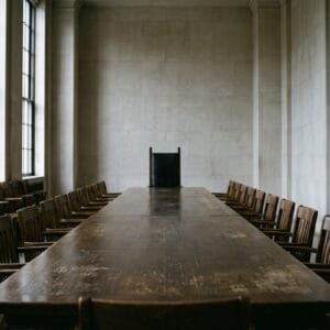 Lange vergadertafel in een sobere zaal met één hoge stoel aan het hoofd en rijen lagere stoelen erlangs