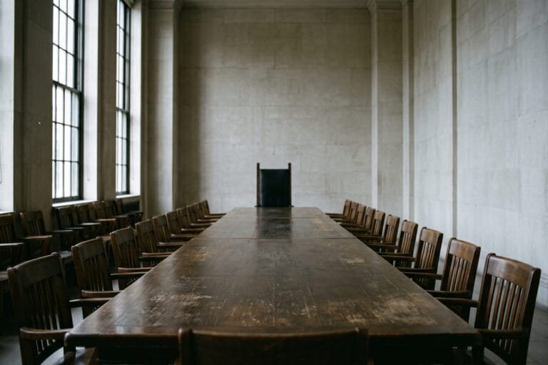 Lange vergadertafel in een sobere zaal met één hoge stoel aan het hoofd en rijen lagere stoelen erlangs