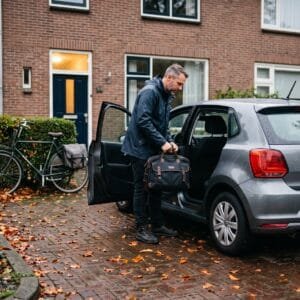 Man zet op een herfstochtend zijn werktas in een kleine auto op de oprit van een rijtjeshuis naast een fiets