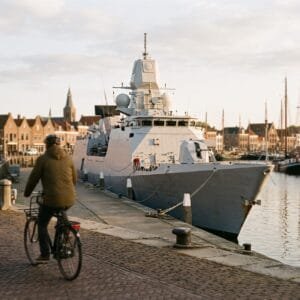 Grijs marineschip ligt in herfstlicht aan de kade van een Nederlandse haven terwijl een fietser voorbijrijdt