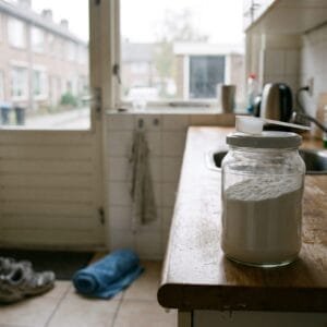 Pot zonder etiket met wit poeder en een maatschepje naast een glas water op een houten aanrecht