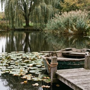 Houten roeiboot aan een steiger in een stille vijver met waterlelies en een treurwilg in de herfst