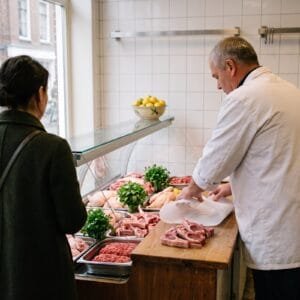 Slager pakt lamskoteletten in papier voor een klant bij een vitrine met lamsvlees, kip en verse kruiden