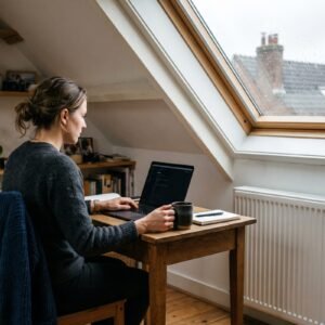 Vrouw werkt met laptop aan een klein bureau op een zolderkamer naast een radiator op een grijze herfstochtend