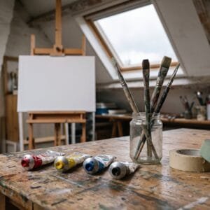 Vier tubes verf in rood, geel, blauw en zwart op een werktafel, met kwasten in een glazen pot en een leeg doek op een ezel